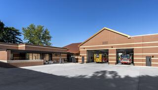 A modern fire station features a brick exterior, two large vehicle bays, and a clear blue sky, surrounded by trees.