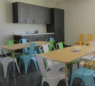 A modern break room features colorful metal chairs around wooden tables, with a kitchenette and dark cabinets in the background.