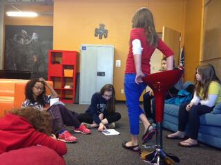 A group of girls interacts in a casual setting, with one standing, while others sit or lie on the floor, engaged in conversation.