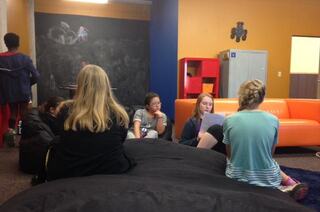 A group of people sits on a large beanbag and around a cozy lounge area with vibrant orange and blue walls, engaged in conversation and activities.