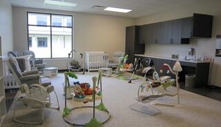 A bright nursery with various baby equipment, including cribs, rockers, and colorful play items, set in a spacious, clean room.