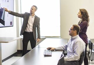 A man in a suit points at a screen while two colleagues observe; one sits at a desk with a keyboard, and the other stands nearby.