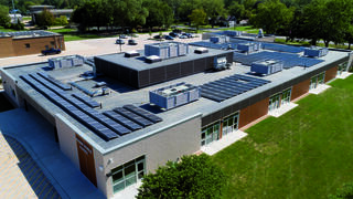 Aerial view of a modern building featuring multiple rooftop units and solar panels, surrounded by green landscape and parking area.