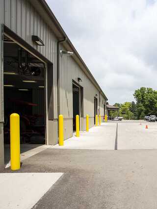 A metal building features open bays and bright yellow safety bollards along a concrete pathway, set against a cloudy sky and green trees.
