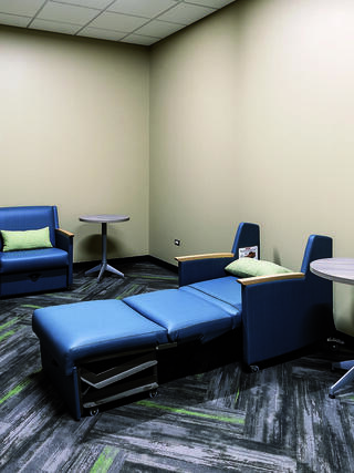 A cozy waiting room features blue seating with green cushions, a recliner, and small tables against a neutral wall and patterned carpet.