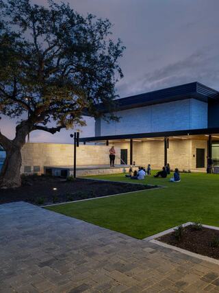 A modern building with a flat roof overlooks a lawn where people gather, enjoying the twilight beneath a large tree.