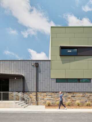A modern building features green and gray panels, with a person walking by and a clear blue sky overhead. Landscaping enhances the scene.