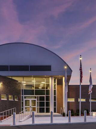 A modern building features a curved roof, large glass entrance, and flags, illuminated against a beautiful twilight sky.