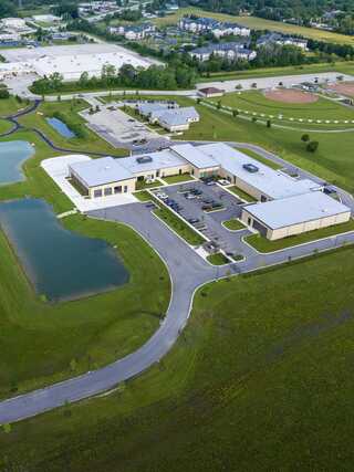 Aerial view of a modern building complex surrounded by green lawns, ponds, and parking, with nearby sports fields and residential areas.