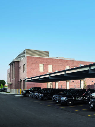 A modern, two-story police station features a row of parked patrol cars under a shaded area, set against a clear blue sky.