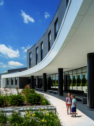 Two children walk along a curved pathway outside a modern building with large windows, surrounded by greenery and under a clear blue sky.