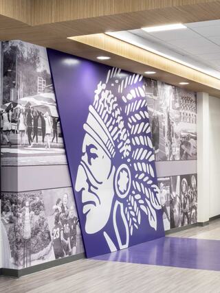 A mural features a large, stylized Native American chief surrounded by historical black-and-white photographs, set in a modern interior.