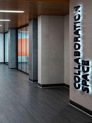 A modern corridor features sleek gray tiles and illuminated signage for "COLLABORATION SPACE," with glass walls revealing vibrant interior colors.
