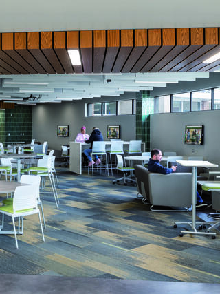 A modern, spacious seating area with tables and chairs, featuring wood accents and large windows, where people engage in conversation and work.