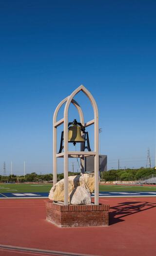 A large bell is mounted on a wooden structure surrounded by large rocks, set against a clear blue sky and a sports field backdrop.