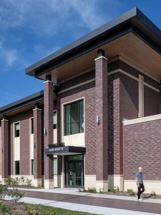 A modern brick building with large windows and a sleek design, set against a clear blue sky. A person walks nearby along a pathway.