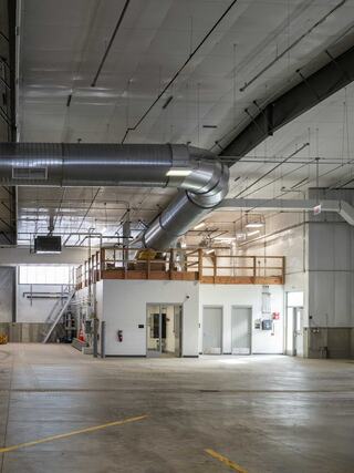 A spacious, industrial interior features a high ceiling, large ventilation ducts, and a mezzanine area with a wooden railing.