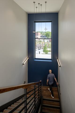 A person descends a modern staircase, illuminated by natural light from a tall window framed in deep blue, against a minimalist backdrop.