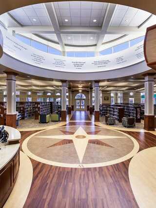 A spacious, well-lit library interior features a circular design with wooden floors, bookshelves, and large windows illuminating the space.