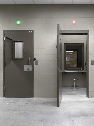 A stark, gray room features two heavy doors, one open. Green and red lights signal activity, with a bench visible in the background.