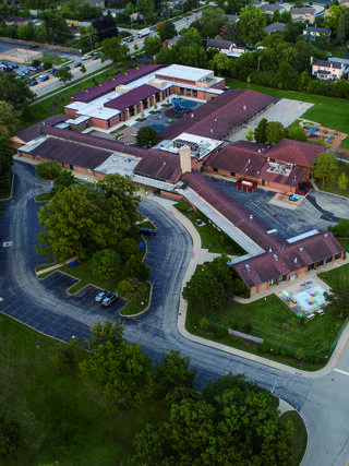 Aerial view of a school complex featuring multiple buildings, a pool area, and green spaces, surrounded by a parking lot and trees.