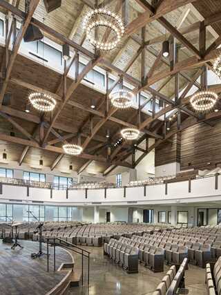 A spacious auditorium features modern architecture with wooden beams, elegant chandeliers, and rows of comfortable seating.
