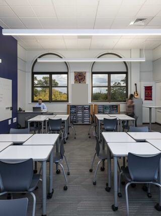A bright, modern classroom features multiple tables, chairs, large windows, and a few students engaged in activities.