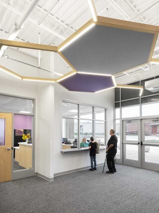 A modern reception area features a hexagonal ceiling design, with a man and child interacting at the reception window. Bright and airy space.