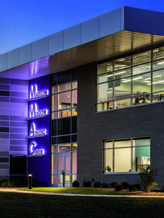 A modern athletic center features sleek architecture and illuminated signage, showcasing its name in vibrant purple lighting at dusk.
