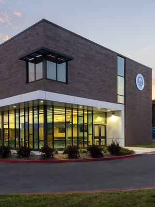 A modern, multi-story brick building features large glass windows and a welcoming entrance, set against a colorful evening sky.