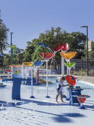 A playful splash pad features colorful water structures, with a child enjoying the water while surrounded by greenery and bright blue skies.