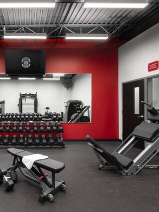A modern gym featuring a weightlifting area with dumbbells, adjustable benches, and ample space, highlighted by red and black decor.