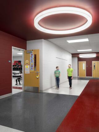 A bright hallway features modern lighting and colorful walls, with two students walking past classroom doors and a welcoming atmosphere.