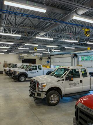 A spacious garage features several white and red trucks parked in neat rows under bright overhead lights, with tools and equipment in the background.