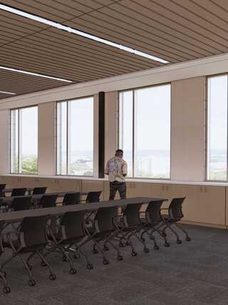 A modern conference room with large windows, featuring a man standing alone, overlooking a cityscape, and surrounded by sleek tables and chairs.