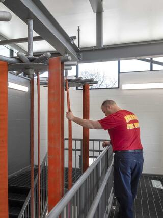 A man in a red shirt inspects fire hoses hanging from a rack in a well-lit, modern fire station with large windows.