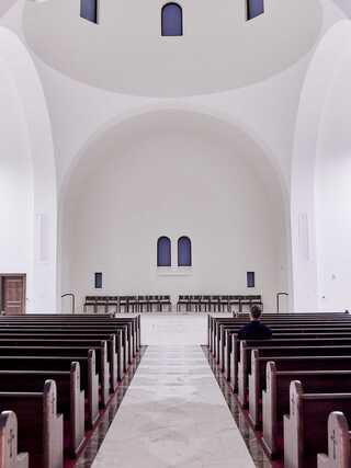 A serene, minimalist interior of a chapel with rows of wooden benches and a lone figure seated, under a high, curved ceiling.