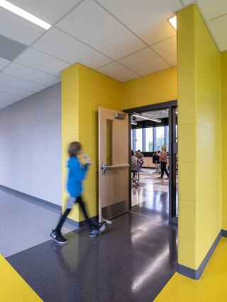 A hallway features a yellow accent wall, an open door leading to a classroom, and students engaged in various activities inside.
