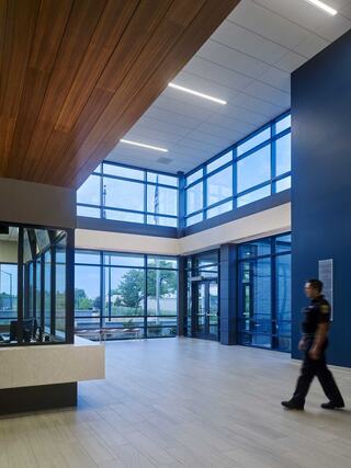 A modern corridor with large glass windows, wooden accents, and a person in uniform walking through the well-lit space.