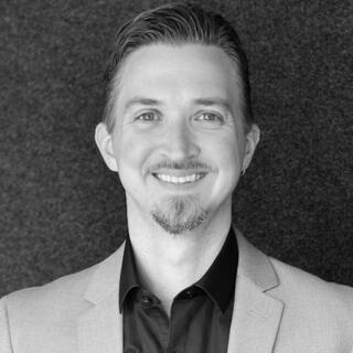 A smiling man with a goatee wears a light blazer over a black shirt, set against a textured dark background.