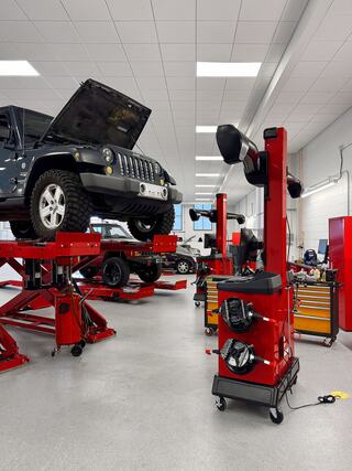 A vehicle is elevated on a lift in a mechanic’s workshop, surrounded by red equipment and tools, with another car visible in the background.