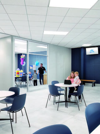 A modern, bright room features tables and chairs. A woman sits with a toddler, while kids play in a glass-walled area nearby.