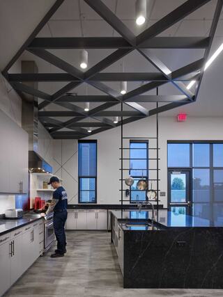 A modern kitchen features sleek cabinets, a large island, and striking geometric ceiling beams, with a chef preparing food.