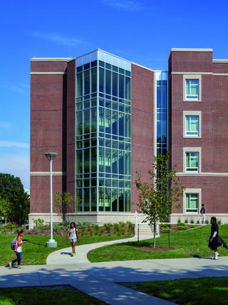 A modern brick building with large glass windows overlooks a landscaped area, while students walk along a pathway. Clear blue sky above.