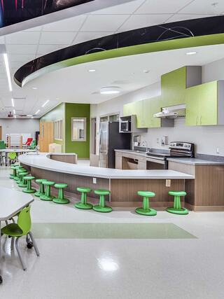 A modern kitchen with a curved bar, green accents, and bright stools, featuring stainless appliances and a spacious, inviting layout.