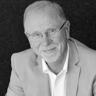 A smiling man with glasses, wearing a light-colored blazer over a white shirt, poses against a dark background.
