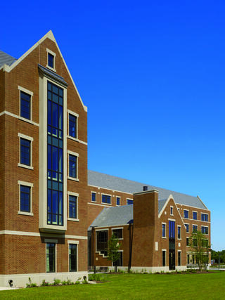 A modern brick building features large windows and a peaked roof against a clear blue sky, surrounded by a grassy area.