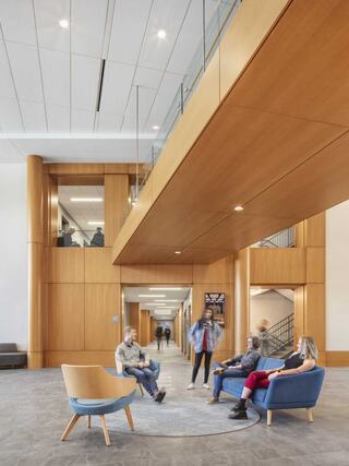 A modern, spacious interior features wooden accents, large windows, and people conversing in a welcoming lounge area.