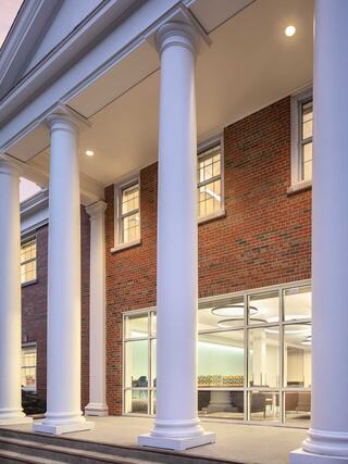 A modern building features large white columns and a mix of brick and glass, showcasing an inviting entrance with warm lighting.