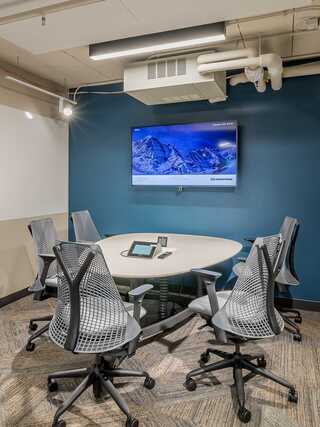 A modern conference room with a round table, four ergonomic chairs, a touchscreen device, and a wall-mounted TV displaying a snowy mountain scene.